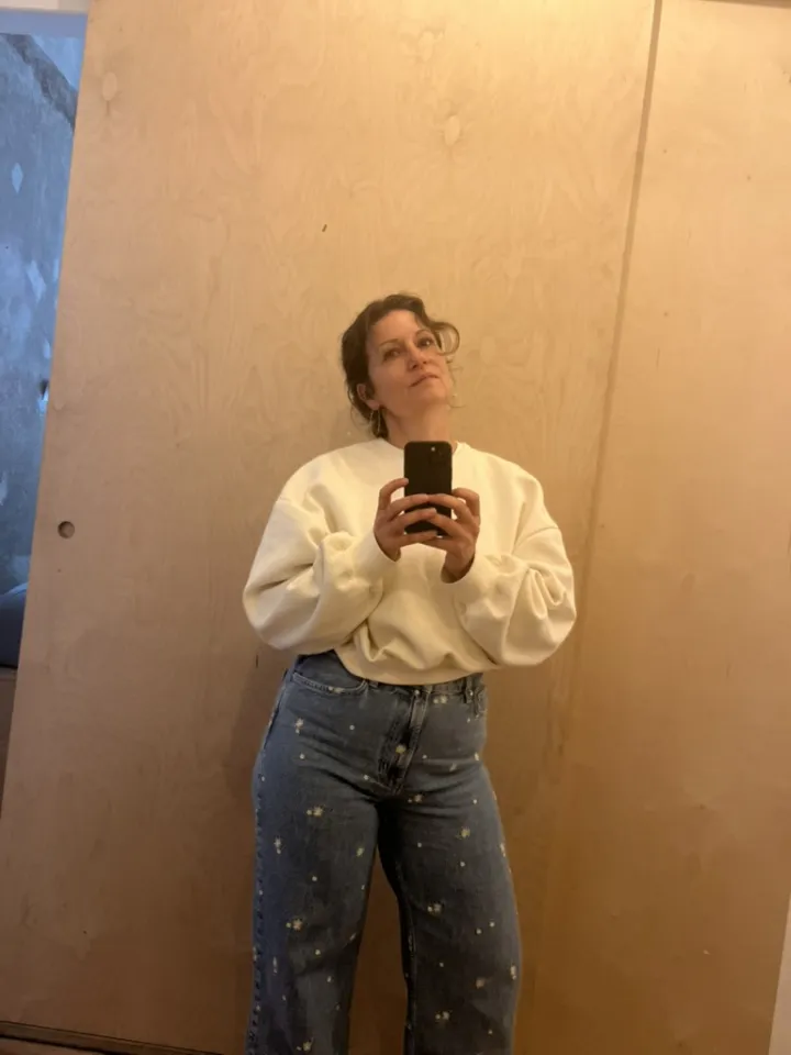 Jenny Gleitsmann in a cream sweater, taking a mirror selfie in front of a light wooden wall